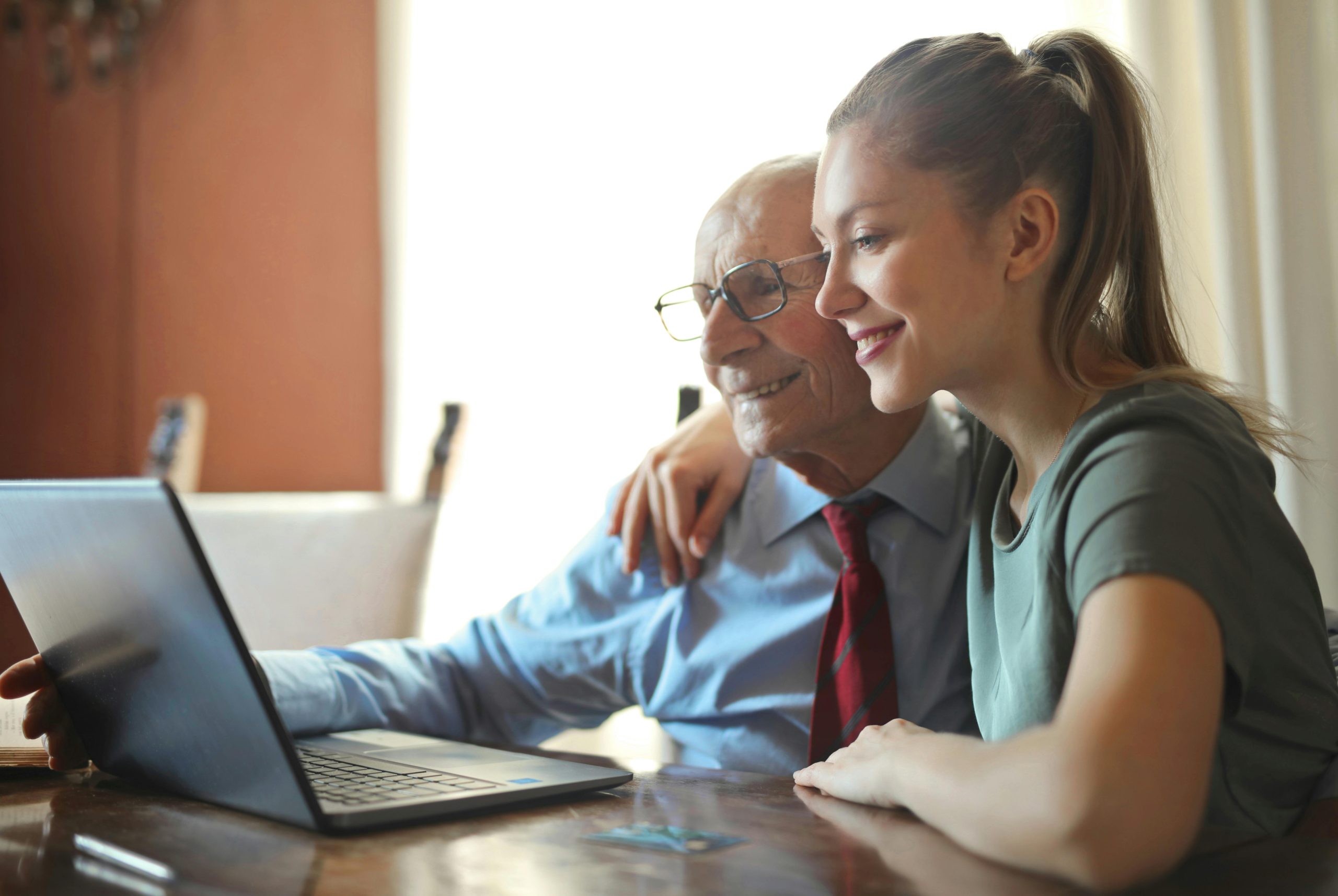 Adult daughter sitting beside her father at a kitchen table reviewing information on a laptop together