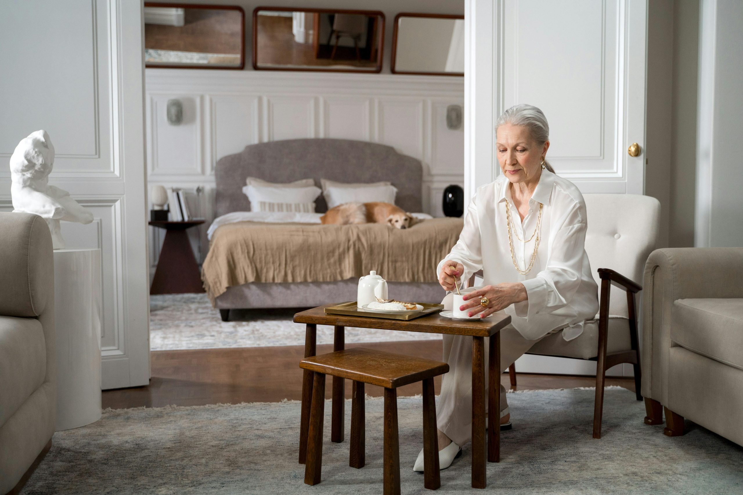 Older woman seated at a small table in her bedroom suite, enjoying tea in a well-lit, comfortable home setting.