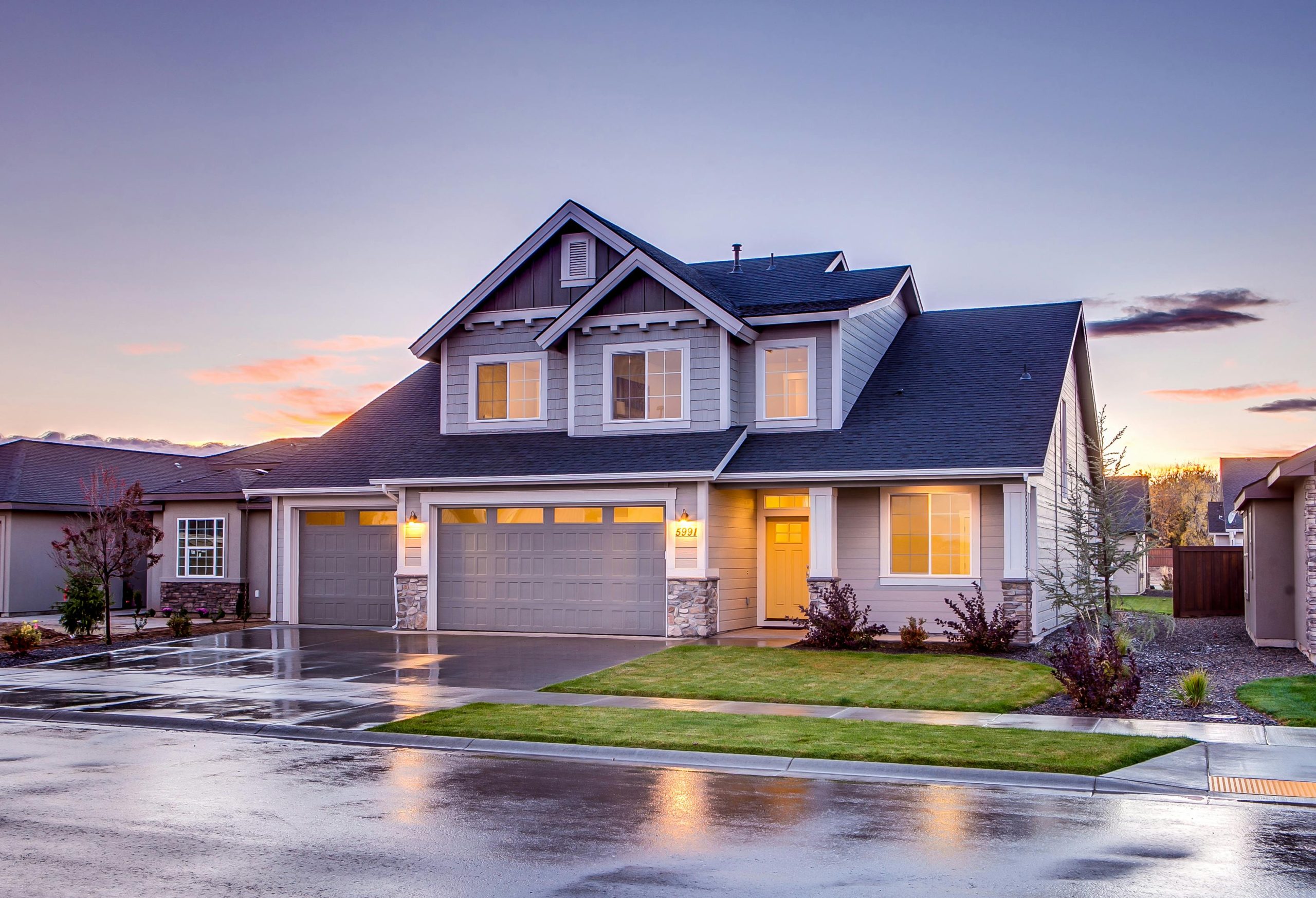 Two-story suburban home at dusk with warm interior lights, landscaped yard, and wet driveway after rain